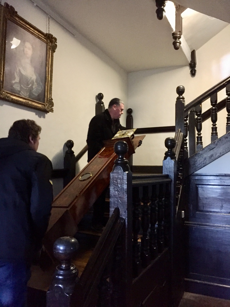 Two technicians Ian and Carl moving a longcase clock with its face exposed up the oak staircase at Aston Hall